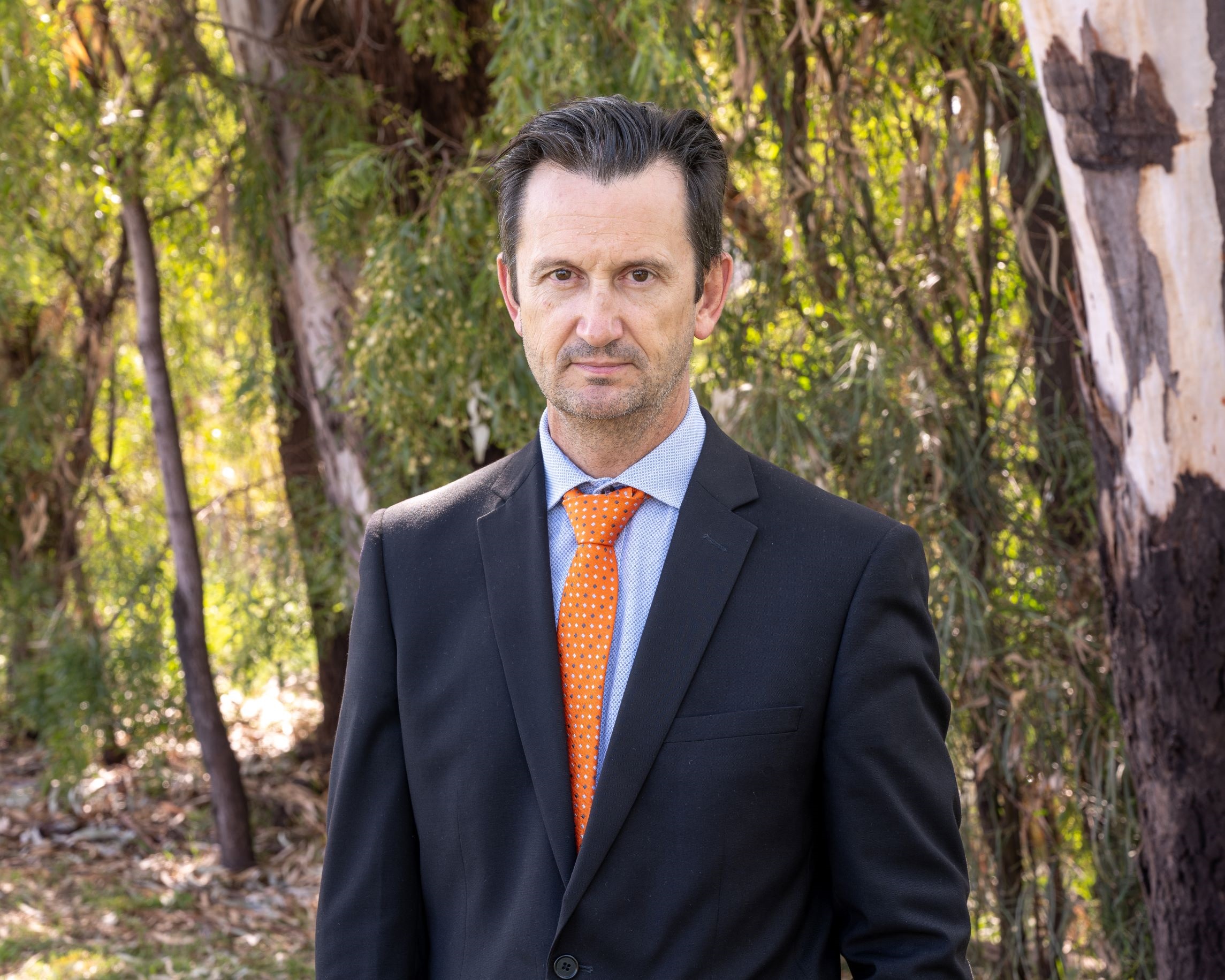 GM Brett Bacon stands in a suit in front of greenery and trees.
