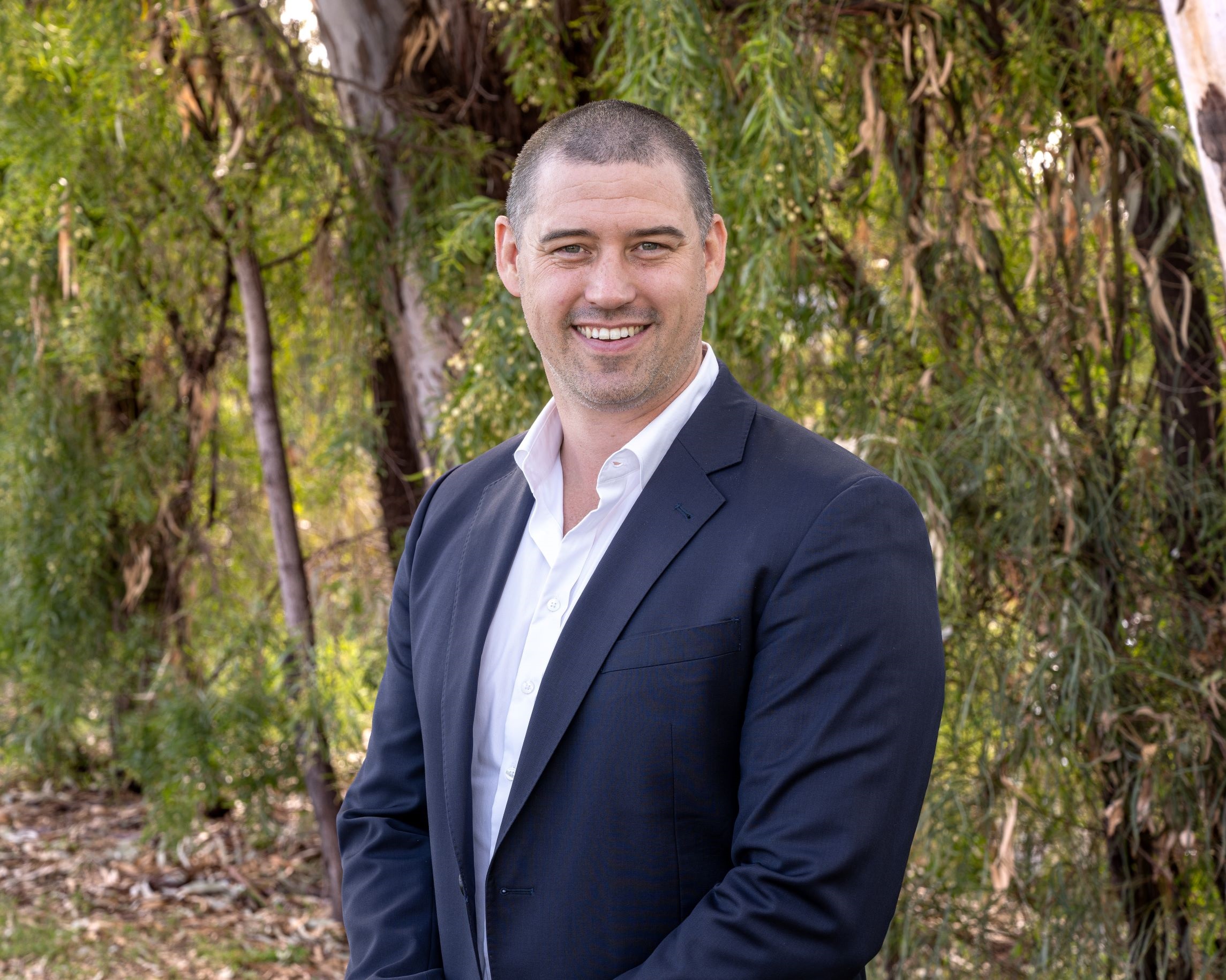 GM Daniel Fletcher stands in front of Myall Creek greenery, wearing a navy suit and smiling.