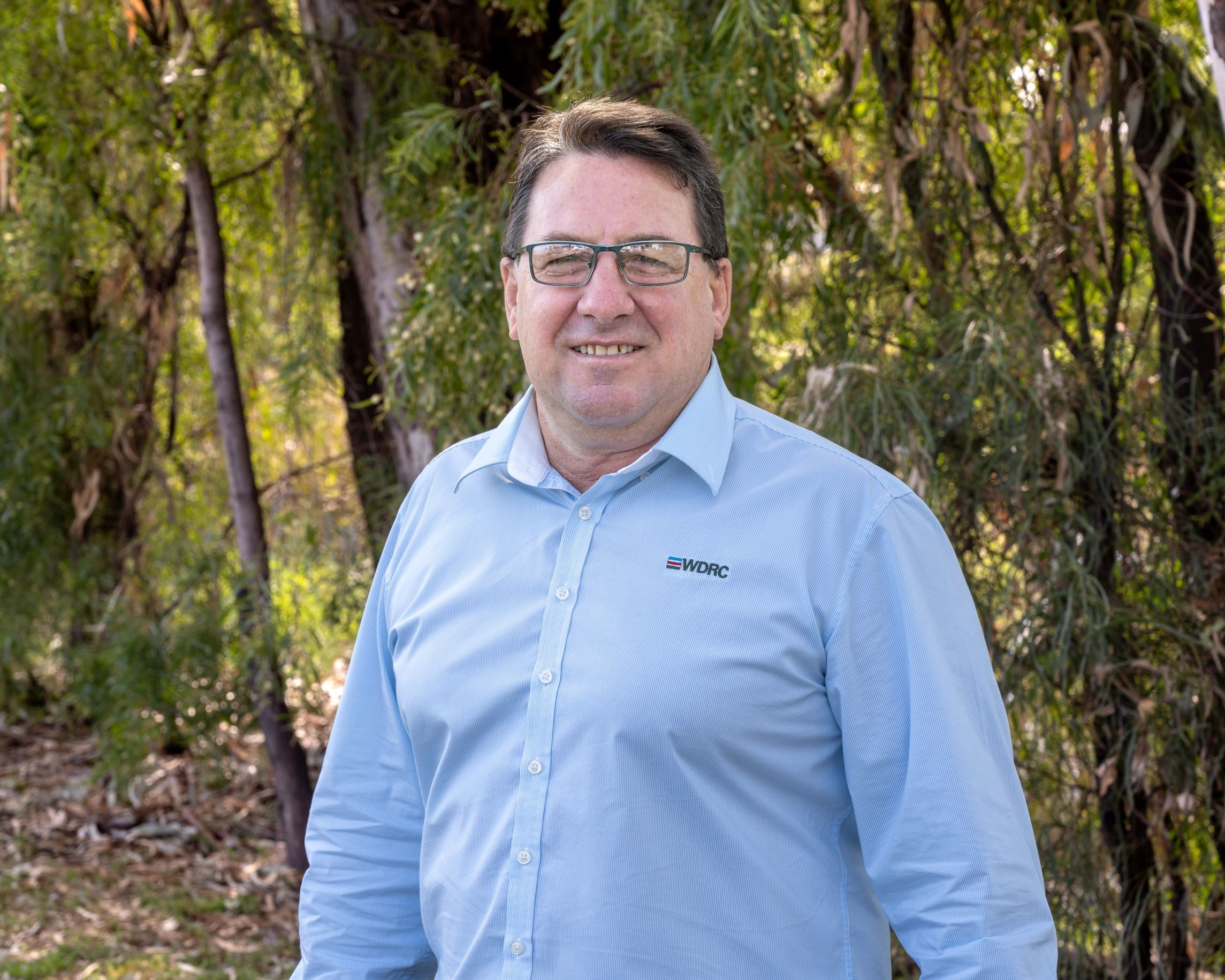 GM Graham Cook stands in a light blue shirt, smiling at the camera, standing in front of Myall Creek greenery.